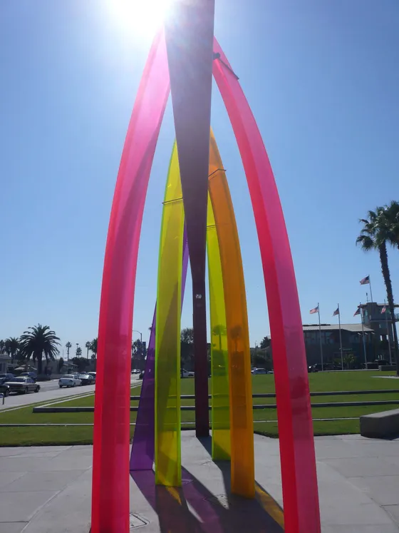 Looking up through Surfhenge at Portwood Pier Plaza: tall acrylic surfboard arches in pink, purple, yellow and orange against a clear blue sky, with palm trees and the Imperial Beach lifeguard tower behind.